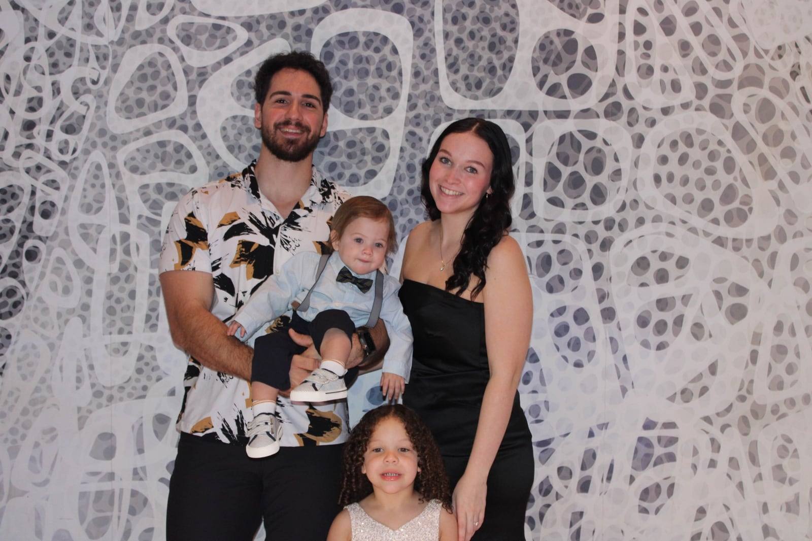 Angelo holding his young son, with his fiancée and his daughter, all smiling in front of a patterned wall.